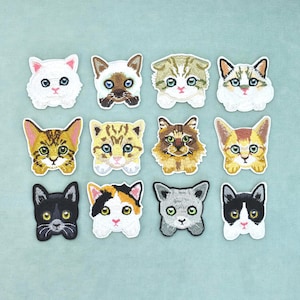 Op de afbeelding: Een verzameling van twaalf geborduurde kattenkop-patches gerangschikt in een raster. De patches tonen verschillende kattenrassen en vachtpatronen, waaronder wit, siamees, cyperse, lapjeskat, zwart en grijs, met gedetailleerd stikwerk en expressieve ogen.