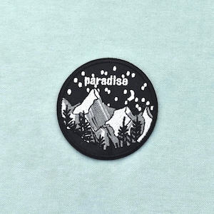 Op de afbeelding: Een ronde geborduurde patch met een zwarte achtergrond, een wit berglandschap, naaldbomen en een sterrenhemel. Het woord paradise is in witte kleine letters boven de bergtoppen geborduurd, omrand door een zwarte cirkelvormige rand.