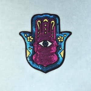 Può includere: Una toppa a forma di mano di Hamsa decorata con paillettes dai colori vivaci magenta, blu brillante e giallo. Il design presenta un motivo a occhio centrale, accenti floreali sui lati e un bordo ricamato nero che delinea la forma della mano.