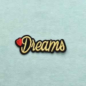 Peut inclure: Un écusson brodé représentant le mot Dreams en écriture cursive jaune avec un contour noir. À gauche du texte se trouve un petit cœur rouge plein. L'écusson est conçu avec un fond noir qui suit la forme des lettres du mot inscrit.
