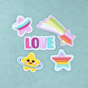 Peut inclure: Un ensemble de cinq écussons brodés comprenant une étoile rose à rayures bleues, une étoile filante avec une traîne arc-en-ciel, le mot LOVE en lettres colorées, une étoile jaune avec un visage et un anneau, et une étoile rayée multicolore.