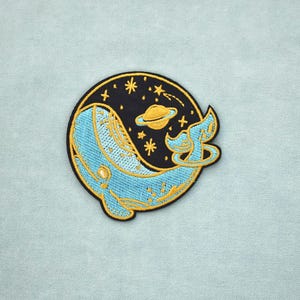 Könnte beinhalten: Gestickter Aufnäher mit einem blauen Wal mit goldenen Akzenten, vor einem schwarzen Hintergrund mit Sternen und einem Planeten. Der Aufnäher hat eine runde Form mit einem goldenen Rand, geeignet für Kleidung oder Accessoires.
