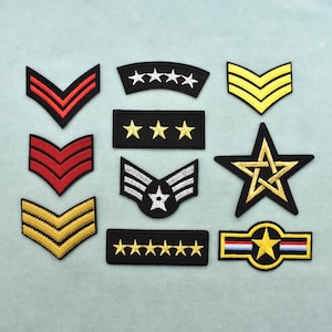 Puede incluir: Una colección de diez parches bordados de estilo militar en varias formas y colores. El conjunto incluye diseños de galones en rojo y dorado, barras rectangulares con estrellas doradas, un emblema de estrella alada, una estrella grande y una barra.