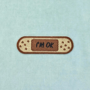May include: An embroidered patch shaped like an adhesive bandage with rounded ends. The centre is a dark brown rectangle featuring the text I'M OK in black thread. The surrounding areas are a light tan colour with small brown dot patterns.