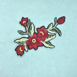 Peut inclure: Un écusson en tissu brodé représentant un groupe de fleurs rouge foncé avec des centres jaunes et des feuilles vert clair. La composition florale est soulignée par une bordure noire épaisse, créant une forme nette sur le fond bleu clair.