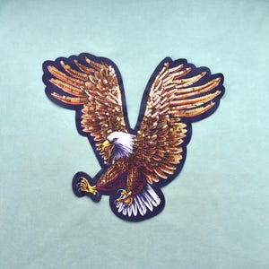 Può includere: Una toppa a forma di aquila con testa bianca, becco e artigli gialli e ali spiegate. Le ali e il corpo sono ricoperti di paillettes dorate e color bronzo scintillanti, creando una superficie strutturata e riflettente su un contorno scuro.
