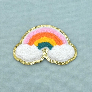 Peut inclure: Un écusson brodé en forme d'arc-en-ciel avec un arc texturé rose, orange, jaune et bleu sarcelle. Le motif comprend deux sections blanches et duveteuses en forme de nuage à la base, le tout bordé d'une bordure dorée scintillante.