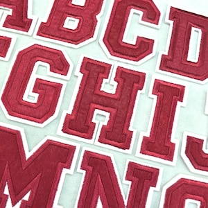 Op de afbeelding: Een verzameling individuele opstrijkbare alfabet-patches met de letters A tot en met T. Elke letter is weergegeven in een vetgedrukt lettertype in collegestijl met een dieprode stoffen kern en een dikke, strakke witte geborduurde rand.