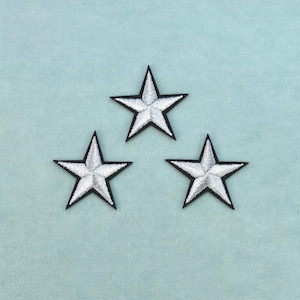 Peut inclure: Trois écussons brodés en forme d'étoile disposés en triangle. Chaque étoile présente un centre en fil argenté métallique avec une bordure noire distincte soulignant la forme à cinq branches, créant un contraste net sur le fond bleu clair.