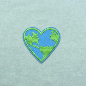 Puede incluir: Un parche bordado en forma de corazón que presenta un fondo azul con un diseño de masa terrestre verde estilizado. El parche tiene un borde azul distintivo y un contorno cosido en verde que sigue la forma del corazón, creando un elemento gráfico detallado.