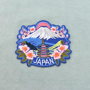 Peut inclure: Écusson brodé représentant un paysage japonais. Le motif comprend une montagne enneigée, une pagode et des fleurs de cerisier. Le mot "JAPAN" est brodé en blanc sur un fond bleu. L'écusson a une bordure bleue.