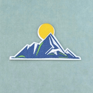 Peut inclure: Un écusson brodé représentant une chaîne de montagnes stylisée en bleu foncé avec des sommets enneigés blancs et de fines touches vertes. Un soleil circulaire jaune vif est placé derrière les sommets. Le motif est bordé de blanc.