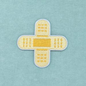 Op de afbeelding: Een geborduurde patch in de vorm van twee kruisende pleisters. De patch heeft een lichtgele kleur met een donkerder geel rechthoekig kussentje in het midden en kleine geborduurde rasterpatronen op de uiteinden van elke arm van het kruis.