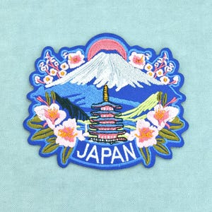 以下が含まれることがあります： 白い雪を頂いた山、ピンクの太陽、多層の塔、そしてピンクの桜が刺繍されたワッペン。デザインは青い縁取りで囲まれており、下部には白い大文字でJAPANという文字が刺繍されています。