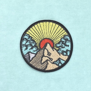 Pode incluir: Um emblema bordado circular apresentando um pico de montanha bege centralizado sob um sol vermelho com raios amarelos. O design inclui nuvens azul-claras estilizadas em ambos os lados da montanha, tudo contornado por uma borda preta espessa.