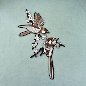 Può includere: Una toppa ricamata raffigurante due uccelli marroni e bianchi appollaiati su rami neri con piccoli fiori bianchi. Un uccello è raffigurato in volo con le ali spiegate, mentre l'altro è mostrato in posizione di riposo con una lunga coda.