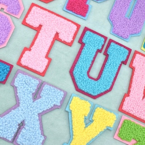 May include: A collection of varsity-style alphabet letter patches featuring a textured, fuzzy chenille surface. Each letter is outlined with a contrasting fabric border in various bright colors including pink, blue, yellow, and green.