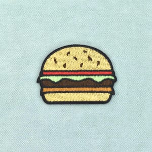 Può includere: Una toppa ricamata a forma di hamburger con un panino color marrone chiaro, una fetta di pomodoro rosso, lattuga verde chiaro, un hamburger di carne marrone e una fetta di formaggio giallo, il tutto bordato di filo nero su tessuto azzurro.