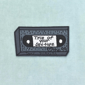Può includere: Una toppa ricamata rettangolare a forma di cassetta audio in tonalità grigie e nere. L'etichetta centrale bianca presenta il testo nero "Title of your sex tape" con "the 99" e un simbolo a forma di cuore sotto, e "JAKE PERALTA" in basso.