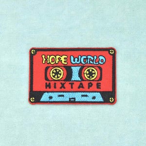 Op de afbeelding: Een rechthoekige geborduurde patch met een rood cassettebandje-ontwerp, zwarte randen en gele schroefdetails. De tekst HOPE WORLD is in geel en blauw geborduurd, met het woord MIXTAPE in het zwart onder de centrale haspels.
