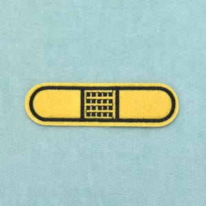 Op de afbeelding: Een rechthoekige pleister-patch met afgeronde uiteinden, voorzien van een felgeel stoffen oppervlak en een centraal zwart geborduurd rasterpatroon. De gehele omtrek van de patch is afgewerkt met een dikke zwarte geborduurde rand.