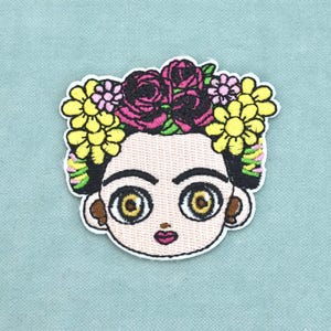 Peut inclure: Un écusson brodé représentant le portrait stylisé d'une personne aux cheveux foncés, aux sourcils épais et aux grands yeux dorés. La figure est ornée d'une couronne vibrante composée de roses rouge foncé, de marguerites jaunes et de petites fleurs roses.