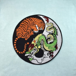 Può includere: Toppa ricamata circolare con un design yin-yang raffigurante una tigre e un drago. La tigre è arancione e nera, il drago è verde, giallo e rosso. La toppa ha uno sfondo nero e un bordo bianco.