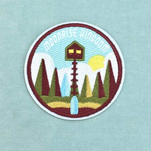 Op de afbeelding: Een ronde geborduurde patch met een landschap van groene en kastanjebruine dennenbomen, een gele zon en een hoge bruine uitkijktoren met gele ramen. De tekst MOONRISE KINGDOM staat in een witte boog bovenaan tegen een lichtblauwe lucht.