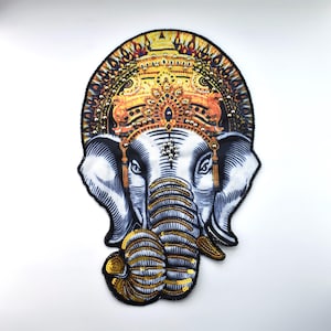 Op de afbeelding: Een geborduurde patch met een olifantenkop met een gedetailleerde kroon en slurf. De patch heeft een wit, zwart, goud en oranje kleurenschema. De kop van de olifant is versierd met pailletten en kralen.