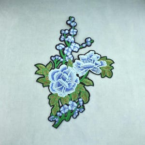 Peut inclure: Un écusson brodé représentant deux grandes fleurs bleu clair semblables à des pivoines, accompagnées de petits boutons bleus sur des tiges vertes avec des feuilles détaillées. L'ensemble est bordé d'un contour noir épais et marqué.