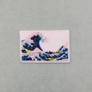 May include: An embroidered iron-on patch featuring a blue and pink wave design, reminiscent of The Great Wave off Kanagawa by Hokusai.