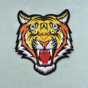 Peut inclure: Écusson brodé tête de tigre avec des détails jaunes, oranges, blancs et noirs. Le tigre a la gueule ouverte avec une langue rouge et des dents acérées. Pour vêtements ou accessoires.