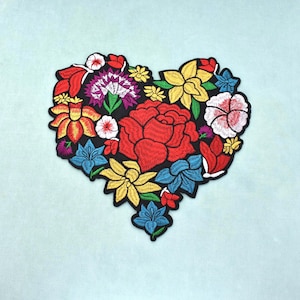 Peut inclure: Un écusson brodé en forme de cœur composé d'un arrangement dense de fleurs colorées, incluant une grande rose rouge centrale, des fleurs jaunes, des lys bleus, ainsi que des touches blanches et violettes, le tout bordé de noir.