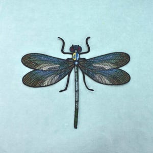 Op de afbeelding: Een geborduurde libelle-patch met iriserende blauwe, groene en zilveren vleugels met zwarte omlijningen. Het lichaam van het insect is gedetailleerd met zwart, blauw en geel garen en is geplaatst tegen een effen lichtblauwe stoffen achtergrond.