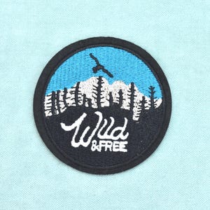 Op de afbeelding: Een ronde geborduurde patch met een zwarte rand. Het ontwerp toont een blauwe lucht, een witte bergtop, zwarte dennenbomen en het silhouet van een zwarte vogel. De witte tekst "Wild & Free" is onderaan op de patch geborduurd.