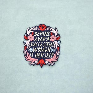 Peut inclure: Écusson brodé avec le texte "BEHIND EVERY SUCCESSFUL WOMAN IS HERSELF" en blanc, entouré d'un motif floral avec des accents roses, rouges et blancs sur fond bleu marine. L'écusson est sur un tissu bleu clair.