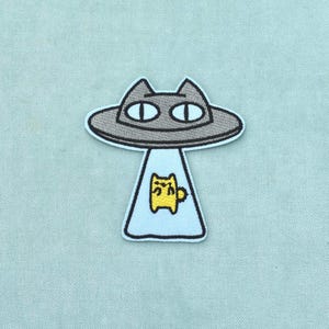 Peut inclure: Un écusson brodé représentant un OVNI en forme de chat gris avec de grands yeux. Un rayon tracteur bleu clair s'étend vers le bas depuis l'engin, contenant une petite créature jaune à quatre pattes avec une queue touffue au centre.
