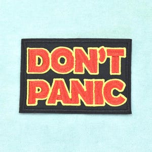 Könnte beinhalten: Ein rechteckiger bestickter Aufnäher mit schwarzem Hintergrund und einem dicken schwarzen Rand. Der Text DON'T PANIC ist in fetten roten Buchstaben mit einer dünnen gelben Umrandung gestickt und in zwei Zeilen mittig angeordnet.