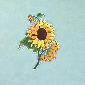 May include: An embroidered patch featuring a large, central sunflower with bright yellow petals and a dark brown centre, accompanied by a smaller sunflower bud, a partially visible flower, and green leaves on a curved green stem.