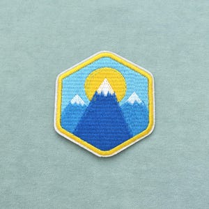 Peut inclure: Un écusson brodé hexagonal représentant un paysage de montagne avec trois sommets bleus enneigés de blanc, sur un ciel bleu clair avec un soleil jaune vif situé derrière le pic central, le tout bordé par un contour de couleur jaune.