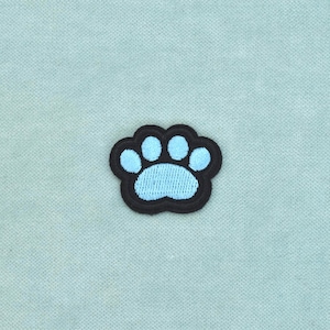 Può includere: Una toppa ricamata a forma di impronta di zampa. Il design presenta un cuscinetto e quattro dita di colore azzurro, il tutto delineato da un bordo nero spesso. Il ricamo presenta un motivo a punti strutturato visibile su tutto il design.