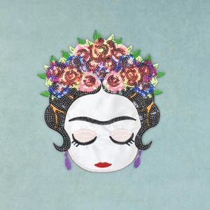 Op de afbeelding: Een decoratieve geborduurde patch met een gestileerd gezicht met gesloten ogen, rode lippen en zwart haar versierd met een levendige kroon van veelkleurige lovertjesbloemen en groene bladeren, geaccentueerd door twee kleine paarse oorbellen.