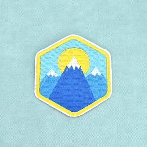 Peut inclure: Un écusson brodé hexagonal représentant une chaîne de montagnes aux sommets enneigés blancs sur un ciel bleu clair. Un soleil jaune vif est placé derrière la montagne centrale. Le motif est entouré d'une bordure jaune épaisse.