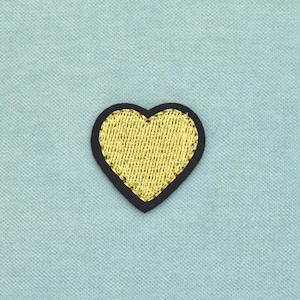 Peut inclure: Un écusson brodé en forme de cœur avec un centre en fil doré métallique et une bordure noire distincte. La broderie présente une texture de fil visible sur toute la section dorée, créant un aspect brillant sur le fond en tissu bleu clair.