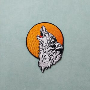 May include: White embroidered wolf howling at a yellow moon. The wolf is facing the right side of the image. The moon is behind the wolf. The image is on a light blue background.