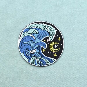 Pode incluir: Um emblema bordado circular apresentando uma onda oceânica azul estilizada a quebrar contra um fundo de céu noturno preto. O design inclui uma lua crescente amarela e várias pequenas estrelas amarelas e brancas espalhadas pelo espaço escuro.