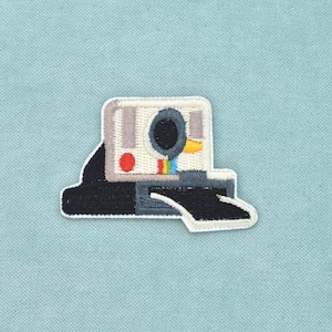 Può includere: Una toppa ricamata a forma di fotocamera istantanea vintage. La fotocamera è bianca, grigia e nera con un pulsante circolare rosso, un accento giallo e un piccolo dettaglio arcobaleno. Una foto rettangolare nera esce dalla fessura frontale.