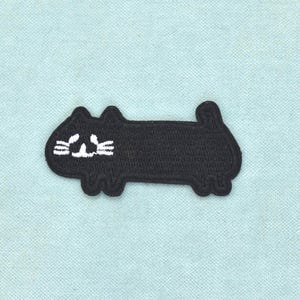 Op de afbeelding: Een zwarte geborduurde patch in de vorm van een lange, rechthoekige kat met een kleine staart en oortjes. De kat heeft eenvoudige witte stiksels voor de ogen, neus en snorharen aan de linkerkant, wat een strakke silhouet van een kat vormt.