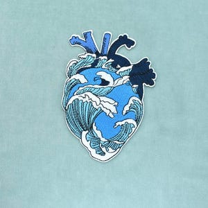 May include: An embroidered patch in the shape of an anatomical heart, featuring a design of crashing ocean waves in shades of light blue, medium blue, and dark navy blue, with white foam accents and a white border outline on a light blue fabric.