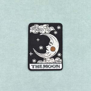 Puede incluir: Un parche bordado rectangular con fondo negro que muestra una luna creciente blanca con rostro, nubes y estrellas. La luna tiene un detalle de mejilla color canela. En la parte inferior, el texto THE MOON aparece en fuente blanca estilizada.